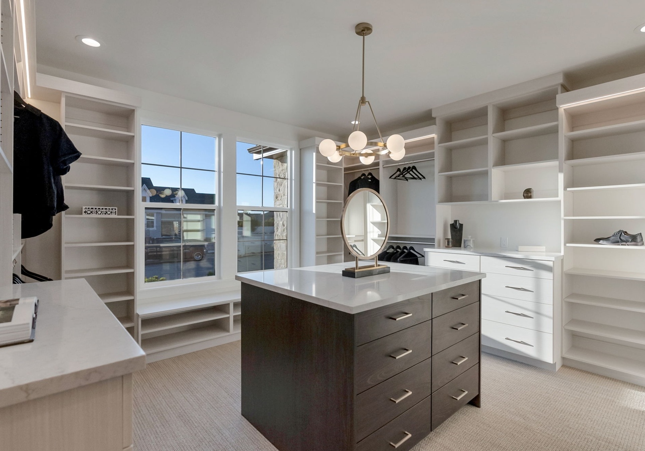 Bright walk-in closet with white built-in shelves, a dark wood island with a marble top, and a round mirror under a modern chandelier.
