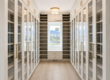 Long, bright walk-in closet with white built-in wardrobes, glass doors, and gold handles along both walls; light wood floor and ceiling lights.