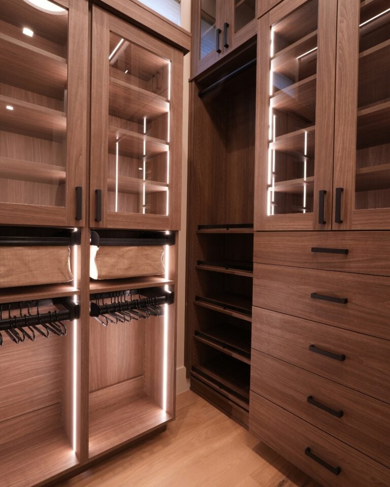 Wooden walk-in closet with glass-front cabinets, open shelves, and a tall dresser, illuminated by integrated LED lighting.