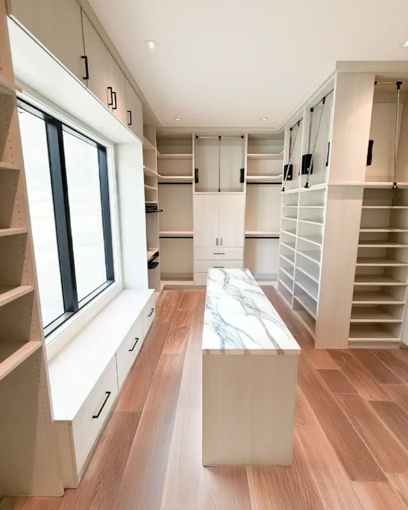 Bright walk‑in closet with white cabinets, open shelves, and a marble‑topped island against wooden flooring.