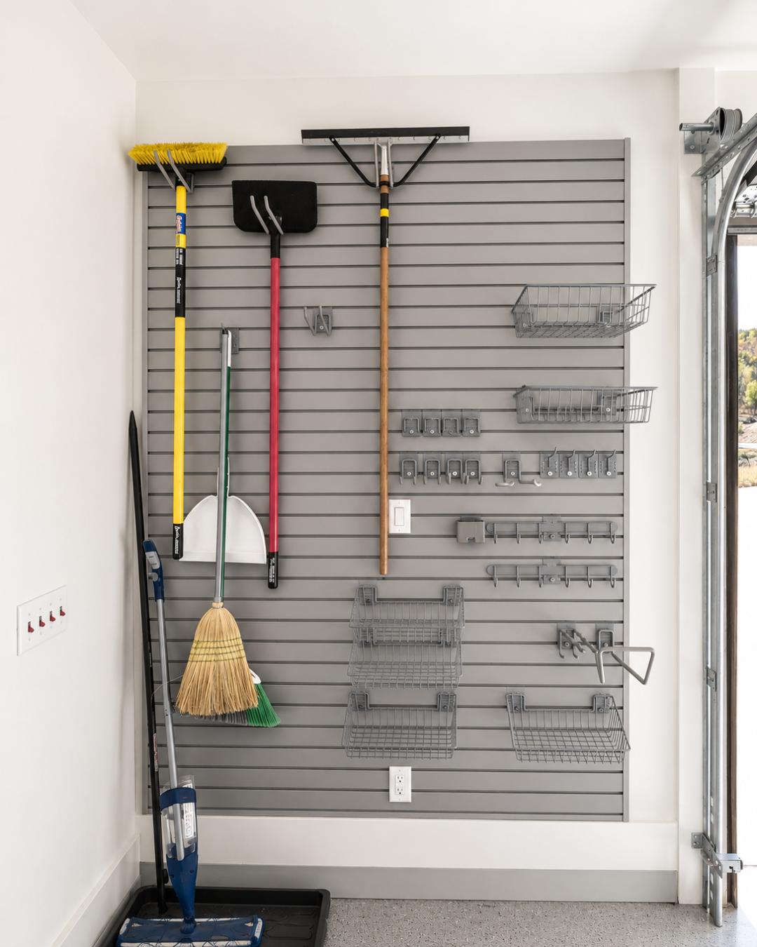 12 Grey pegboard wall in a garage with cleaning tools: broom, rake, shovel, dustpan, and metal baskets.